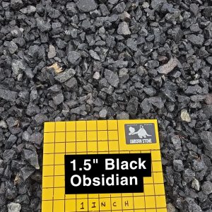 A collection of small black obsidian rocks scattered on the ground. A yellow grid paper labeled "1.5'' Black Obsidian" and "1 Inch Squares" is placed among the stones, indicating the size of the rocks.