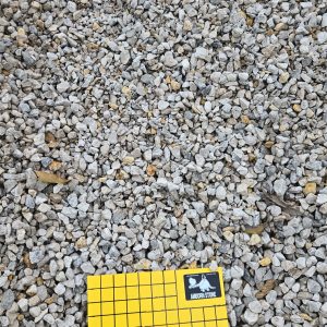 A square yellow grid paper labeled "3/4 clean" lies on a gravel surface. The grid has a small card in the top right corner. The gravel consists of small gray and beige stones.