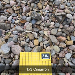 A variety of smooth, multicolored stones are spread across the ground. In the foreground, there's a yellow plastic sample tag with a grid pattern displaying the text "1x3 Cimarron" and "1 inch squares.
