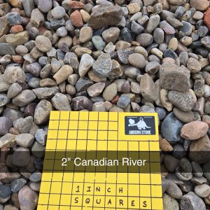 A pile of smooth, multicolored river stones with a yellow grid sign labeled "2'' Canadian River," indicating 1-inch squares for scale. The stones vary in shades of gray, brown, and beige.