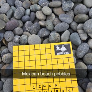 A square yellow grid labeled "1 inch squares" with a sticker reading "Amador Stone" is placed on smooth, round pebbles labeled "Mexican beach pebbles." The pebbles vary in shades of gray and blue.