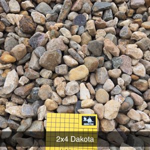 A large pile of variously sized and colored rocks with a yellow grid-patterned sign that reads "2x4 Dakota" and "1/2 inch squares" placed among them.