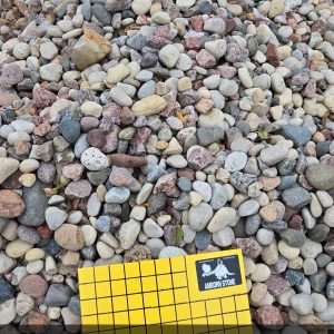 A pile of mixed color, smooth stones with a yellow grid chart labeled "1 Inch Squares" in the foreground. A label reads "1.5" Aspen" on the chart.