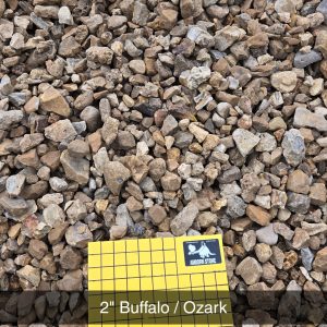 A pile of small, rough stones in shades of brown and gray. In the center, a yellow square grid card is labeled "2'' Buffalo / Ozark" with "1 inch squares" written below.