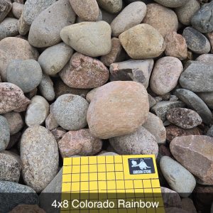 A collection of smooth, multicolored river rocks. In the foreground, there's a cardboard label reading "4x8 Colorado Rainbow, 1 Inch Squares," resting on the stones.