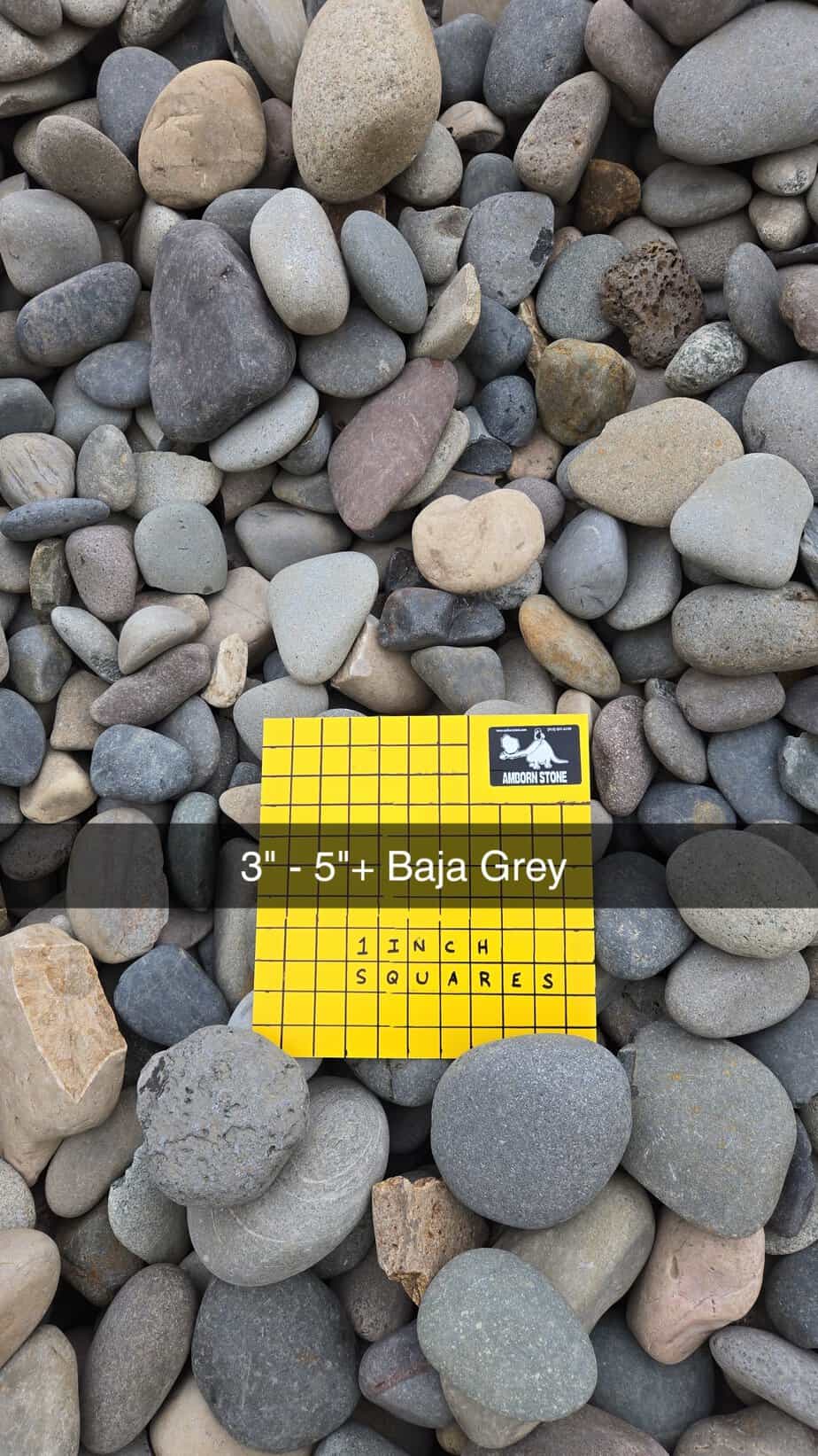 A variety of smooth, round stones in shades of grey and beige. A yellow square grid paper labeled "3'' - 5''+ Baja Grey" and "1 inch squares" is placed on top for size reference.