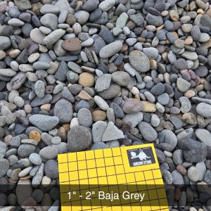 A collection of smooth, rounded grey stones scattered on the ground. A yellow grid card with "1 inch squares" and "1" - 2" Baja Grey" text is partially visible in the foreground.
