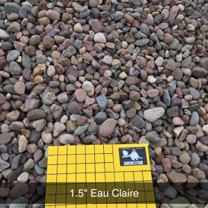 A pile of multicolored small stones with a yellow grid for scale. The grid reads "1 INCH SQUARES" and carries a label "1.5'' Eau Claire Amidon Stone".