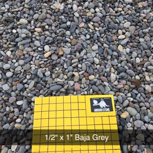 A pile of small, rounded stones with a sample display in front labeled "1/2'' x 1'' Baja Grey" on a yellow grid. The grid has "1 INCH SQUARES" written on it.
