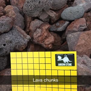 A collection of reddish-brown lava chunks is displayed in a metal cage. A yellow grid card marked "1 inch squares" is placed on top, showing the size of the rocks. The card also features a black and white Amborn Stone logo.