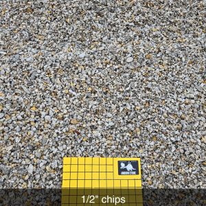 Close-up of small gravel chips with a yellow grid board labeled "1/2'' chips" and "1 inch squares" at the bottom center. The grid board features a logo in the top right corner.