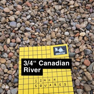 A label reading "3/4'' Canadian River" and "1 Inch Squares" is placed on a bed of multicolored pebbles. The label is yellow with a grid pattern.