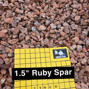 A pile of reddish gravel stones is displayed. A yellow grid board with "1.5'' Ruby Spar" and "1 inch squares" written on it rests on top, showing stone size comparison.