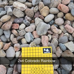 A collection of multicolored river stones spread out, with a yellow gridded card labeled "2x4 Colorado Rainbow" and "1 inch squares" placed on top of them.